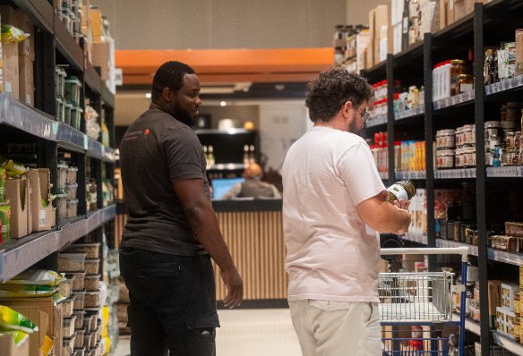 Een medewerker van mdh foodservice in een grijs shirt helpt een klant die naar producten op een schap in het supermarktgangpad kijkt.