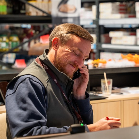 Homme assis à un comptoir, souriant tout en téléphonant et en prenant des notes. À l’arrière-plan, des étagères remplies de produits.