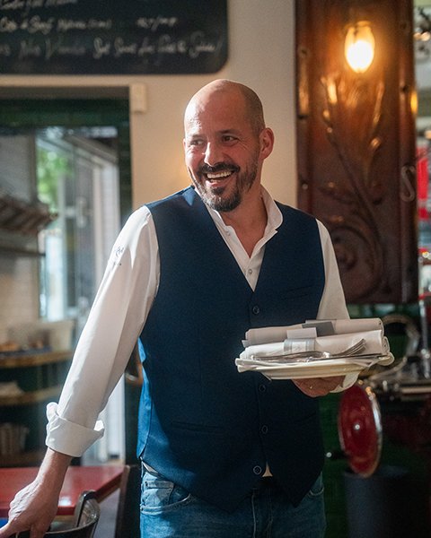 Een portret van Manuel Antonio Rodrigues, een lachende man met een baard, die een gilet draagt en borden vasthoudt in een restaurant.