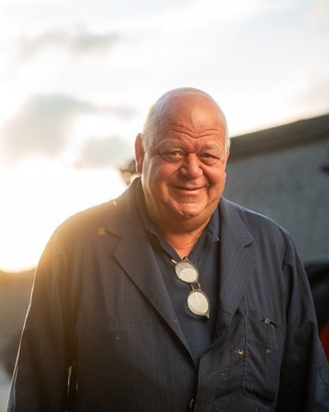 Een portret van Patrick Vanderhaege, een lachende man van middelbare leeftijd in een donker werkhemd, buiten gefotografeerd.